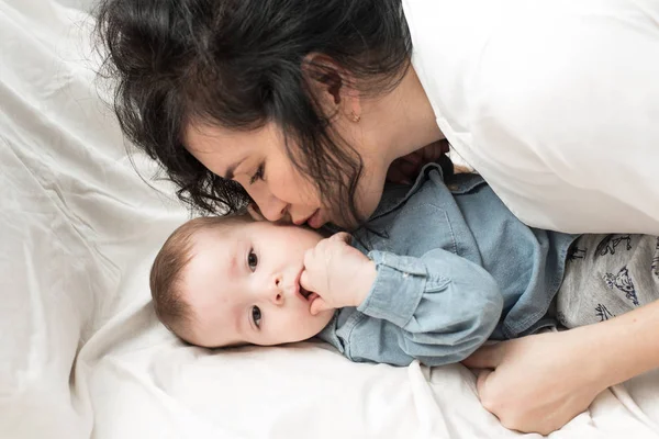 Die dunkelhaarige Mutter küsst ein einjähriges Baby in einem hellen Bett. Mamas Liebe zu ihrem Sohn — Stockfoto