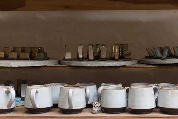 glazed ceramic dishes stand on a shelf in the workshop shelving with handmade ceramic and clay mugs