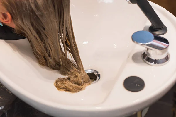 Lavarse el pelo en la cabaña. una peluquera lava el cabello con un champú a una chica rubia . — Foto de Stock