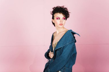 Samimi bir geniş gülümseme ile bir kız bir pastel pembe arka plan üzerinde stüdyoda duruyor. Garip uçan saç modeli ve parlak makyaj ile bir kız