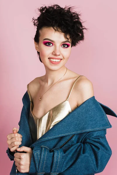 Een meisje met een oprechte brede glimlach staat in de studio op een pastel roze achtergrond. Een meisje met een vreemd vliegende kapsel en heldere make-up. — Stockfoto