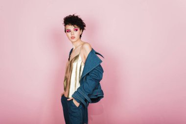 Samimi bir geniş gülümseme ile bir kız bir pastel pembe arka plan üzerinde stüdyoda duruyor. Garip uçan saç modeli ve parlak makyaj ile bir kız