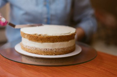 Fırında pasta yapan sevimli bir kız. Kız pastanın üzerindeki kremayı yumuşa. Ahşap bir masada beyaz kek.