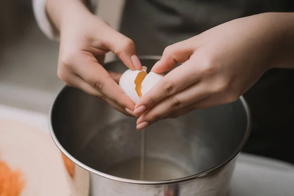 Женщина-повар в сером фартуке разбивает яйцо в металлической чаше. Отделить белок от желтка — стоковое фото