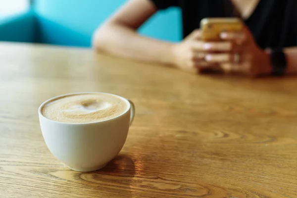 Cappuccino coffee with milk froth on a wooden table. A coffee mug in a coffee shop next to a girl sitting on the phone.