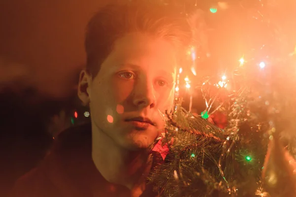 一个年轻的青少年坐在新年树的背景与花环和闪亮的圣诞球。年轻人的新年画像 — 图库照片