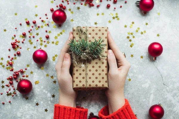 Regalo artesanal de papel marrón con ramitas de árbol de Navidad y cordel en las manos de una niña en un suéter rojo. Regalo hecho a mano sobre un fondo gris grange con juguetes rojos de Navidad en las manos de una chica con —  Fotos de Stock