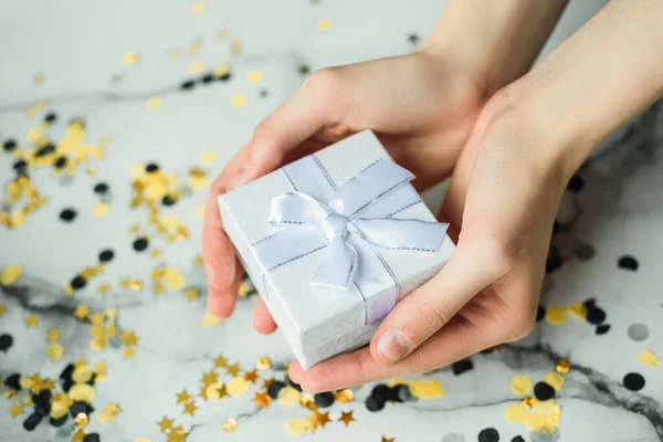 Presente de prata nas mãos de uma menina sem manicure em um fundo cinza com ouro e confete preto. Conceito do Ano Novo e Natal. Vista lateral — Fotografia de Stock