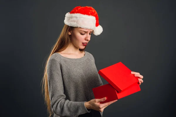 Linda rubia en el sombrero de Santa abre un regalo de Año Nuevo rojo para Navidad. Insatisfecho con el regalo, no hay alegría en recibir la víspera de Año Nuevo en la caja —  Fotos de Stock