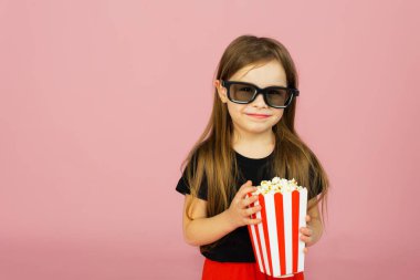 Sevimli küçük kız çizgili ambalajdan patlamış mısır yiyor ve 3D gözlüklü bir film izliyor. Sinema reklamcılığı konsepti, çevrimiçi film izlemek.