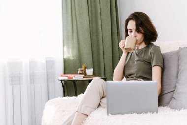 Yatak odasındaki dizüstü bilgisayarla bir fincan sıcak kahve ya da çay içen genç bir kadın. Kadın sosyal uygulamaları kontrol ediyor, online alışveriş yapıyor ve çalışıyor. İletişim ve teknoloji kavramı.