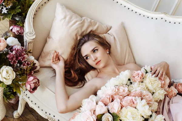 Estilo de boda. Retrato de una hermosa novia en un elegante vestido blanco de rosas, peonías y flores, se encuentra en un sofá en el interior — Foto de Stock
