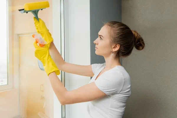 Menina adulta bonito lava uma janela com um raspador. Limpeza de primavera. Um funcionário de uma empresa de limpeza limpa janelas de plástico na casa — Fotografia de Stock