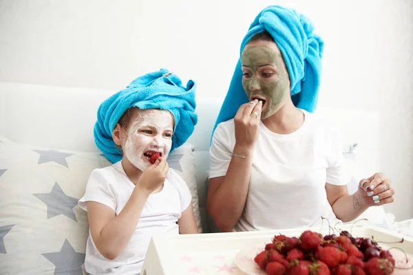 Cute mama i jej córka w masce twarzy piękno jeść truskawki siedzi na łóżku. Rutyna piękna — Zdjęcie stockowe