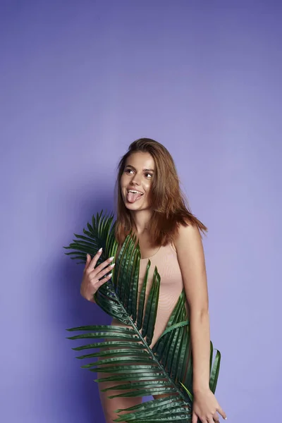 The model smiles and rejoices, has fun and is surprised. A beautiful girl with a tropical leaf on a purple background shows faces and tongue. Funny portrait