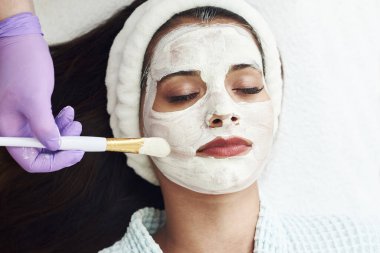 Spa konsepti. Güzellik salonunda besin maskesi takan genç bir kadın, yakın plan. Güzellik uzmanı müşterinin yüzüne nemlendirici bir maske takar..