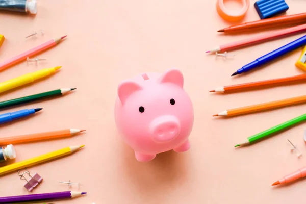 saving for education or back to school concept, piggy bank and colorful marker, pencil and pen on pink background.