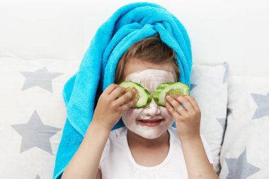 Sabah bakım rutini. Küçük bir kız yüzüne kilden bir maske takıyor ve gözlerinin önünde salatalık kupaları tutuyor..