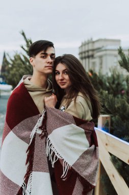 Güzel genç bir çift, şehir caddesinde bir Noel ağacı pazarının arka planında sıcak bir battaniyeye sarılır..