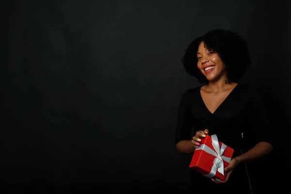 A black woman in a festive black dress holds a Christmas red present with a white ribbon in her hands. The concept of giving or receiving gifts for the new year.