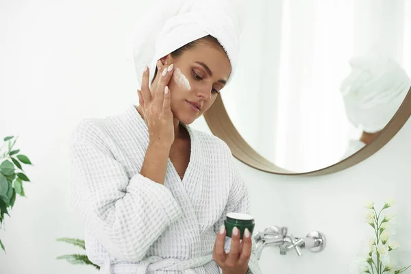 Schöne junge Frau cremt sich im Badezimmer ein. Gesichtscreme. Hautpflege. Schöne lächelnde Frau mit einem Handtuch auf dem Kopf nach dem Duschen — Stockfoto