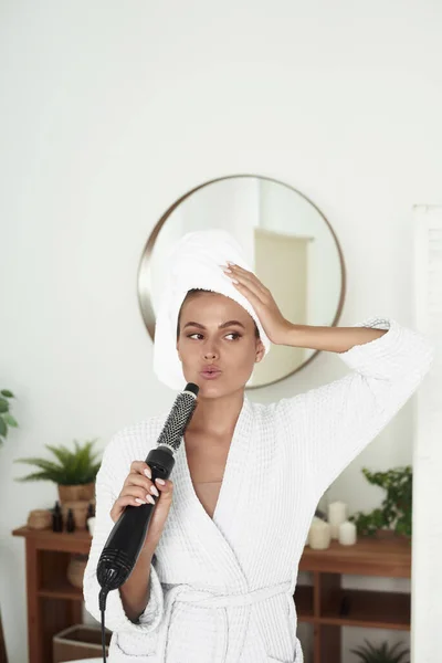 Mujer joven expresiva en albornoz y cabello envuelto en una toalla utiliza un secador de pelo como micrófono — Foto de Stock
