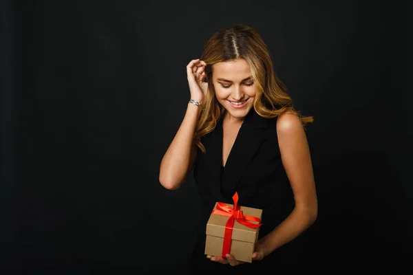 Una encantadora rubia sobre un fondo negro sostiene un regalo artesanal con una cinta roja. El concepto de comprar regalos el viernes negro para Navidad y año nuevo —  Fotos de Stock