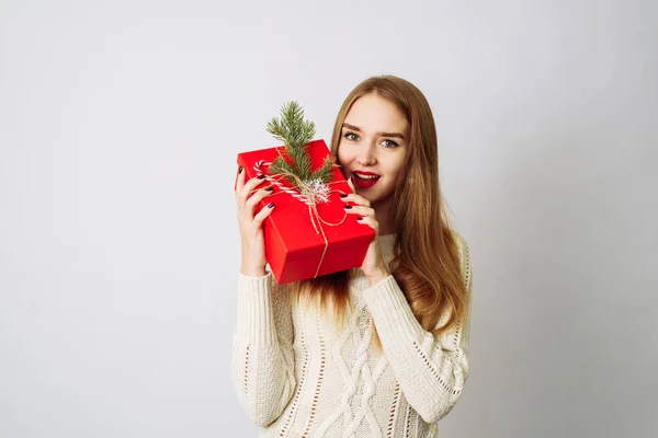 Jovem surpresa segura em uma camisola branca segurando um presente vermelho de Natal com um ramo de abeto e um arco festivo. O modelo feliz está feliz por receber um presente e abre a boca de surpresa — Fotografia de Stock