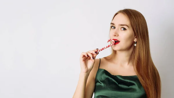 Linda mujer joven en vestido de Navidad de seda verde está mordiendo un bastón de caramelo sobre un fondo blanco — Foto de Stock