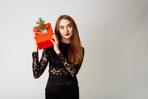 Surprised young woman holds in a black dress holds a Christmas red gift with a fir-tree branch and a festive bow. The happy model is happy to receive a gift and opens her mouth in surprise