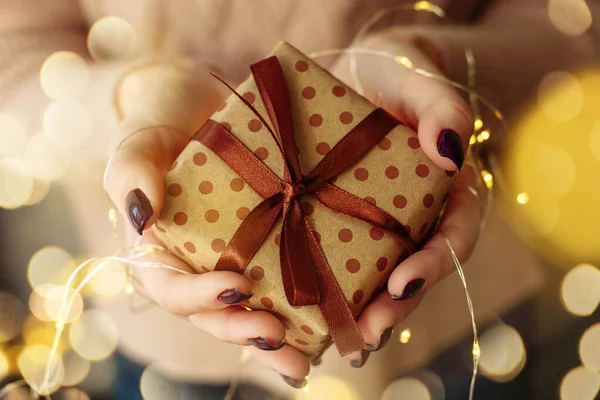 Mãos segurar e dar um presente de embalagem marrom artesanal. Uma menina em uma camisola de malha rosa mantém um Ano Novo, presente de Natal com uma guirlanda com luzes amarelas — Fotografia de Stock