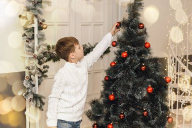 Sevimli küçük çocuk Noel ağacını yeni yıl oyuncakları ve kırmızı toplarla süslüyor. Beyaz örgü kazaklı bir çocuk hediyelerin yanına oturur ve yapay bir ladin üzerine taşak asar. Aile Noel konsepti