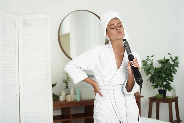 Jovem expressiva em roupão de banho e cabelo envolto em uma toalha usa um secador de cabelo como microfone — Fotografia de Stock