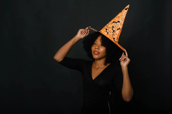 Colpo orizzontale di una giovane donna nera gioiosa con taglio di capelli afro che indossa un cappello arancione di Halloween e un vestito nero. Studio ritratto su sfondo nero — Foto Stock