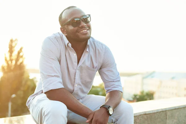 A dark-skinned guy in sunglasses sits in the park. Portrait of a late 20s black man sitting at the park on a summer day with filtered effect — Stock Photo, Image