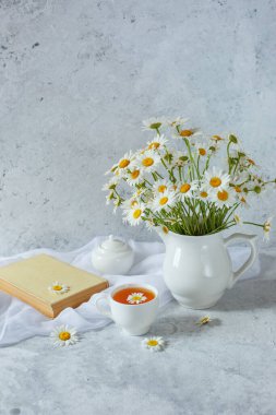 Hayat dolu bir vazo, papatyalar bir fincan çay ve arka planda eski bir kitap.