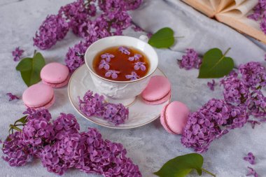 Üzerinde leylak çiçekleri ve badem kurabiyeleri olan bir fincan çay gri arka planda yatar, günaydın yazılı bir kartpostal.