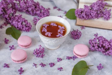Üzerinde leylak çiçekleri ve badem kurabiyeleri olan bir fincan çay gri arka planda yatar, günaydın yazılı bir kartpostal.