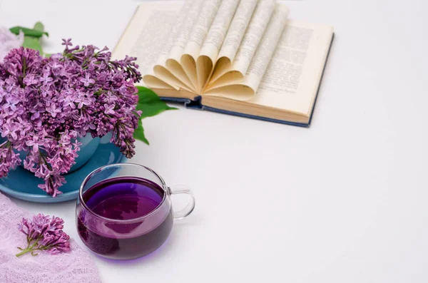 A cup of fragrant purple Clitoria tea and a bouquet of lilac in a blue cup, an open book on a white background. Selective focus, romantic concept, horizontal background, copy space