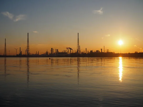 La silhouette d'une raffinerie de pétrole près de l'eau au lever du soleil . — Photo