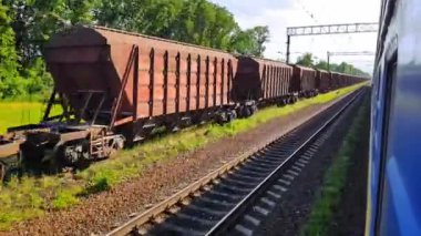 Yolcu treninin yüksek hızlı görünümü yük treninden uzaklaşıyor. Arabanın penceresinden manzara, otobüs, tren. Güneşli bir günde trenden yolculuk
