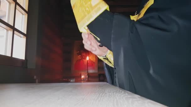 Православный священник надевает священную одежду, стоящую у окна у алтаря деревянной церкви в сумерках — стоковое видео