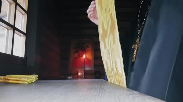 Православный священник надевает священную одежду, стоящую у окна у алтаря деревянной церкви в сумерках — стоковое видео