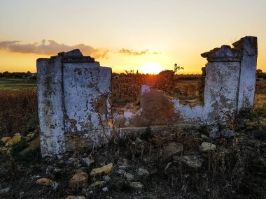 Gün batımı arka planıyla yıkılmış eski kırsal ev. Batan güneşin ışınları sonbaharın başlarında devedikeni çalılıklarında eski bir çiftlik evinin kalıntıları arasında parlıyor.