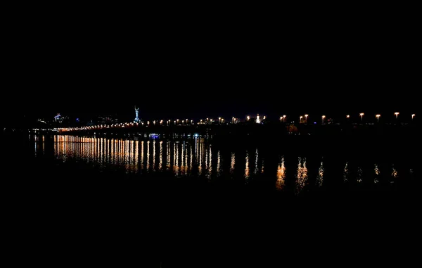Widok nocny mostu Paton przez szeroki Dniepr i światła prawego brzegu Kijowa. Latarnie długiego mostu odbijają się w szerokiej wodzie Dniepru w nocy. — Zdjęcie stockowe