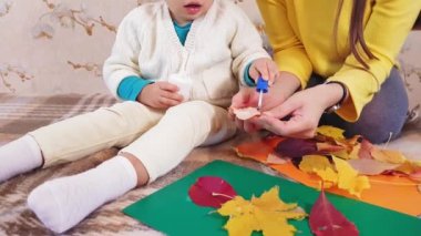 Anne ve küçük kızı güz yapraklarından sonbahar uygulaması yapıyorlar. Annesi olan küçük bir kız, sonbahar koleksiyonunu karton bir çarşafa yapıştırıyor. Evde sonbahar uygulaması yapmak.