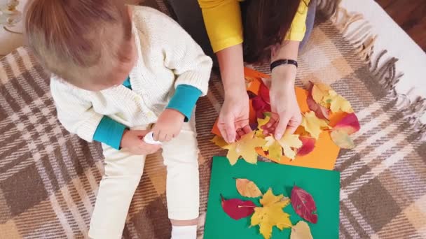 Η μαμά και η μικρή κόρη κάνουν φθινοπωρινή απλικέ από φύλλα φθινοπώρου. Κοριτσάκι με μαμά κολλά φθινοπωρινή ρίψη σε χαρτόκουτο. Κάνοντας το φθινόπωρο απλικέ στο σπίτι. — Αρχείο Βίντεο