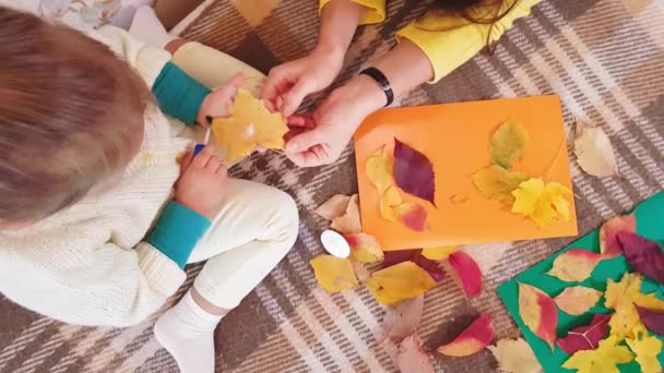 A mãe e a filha pequena fazem o aplique de outono de folhas de outono. Menina com mãe cola fundição de outono em uma folha de papelão. Fazendo aplique outono em casa . — Vídeo de Stock