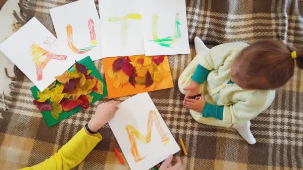年轻的母亲和女儿在白纸上画了一幅写着"秋天"的油画，画了一幅贴画。 小女孩和妈妈一起在白纸上画了一个写着秋天字样的油画. — 图库视频影像