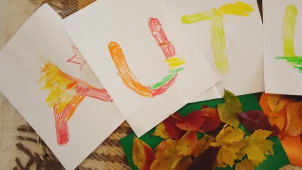 Joven madre e hija dibujan la inscripción pasteles al óleo Otoño en hojas blancas de papel y hacen un apliques. Niña con mamá dibujar letras de aceite Inscripción de otoño en hojas blancas de papel . — Vídeos de Stock